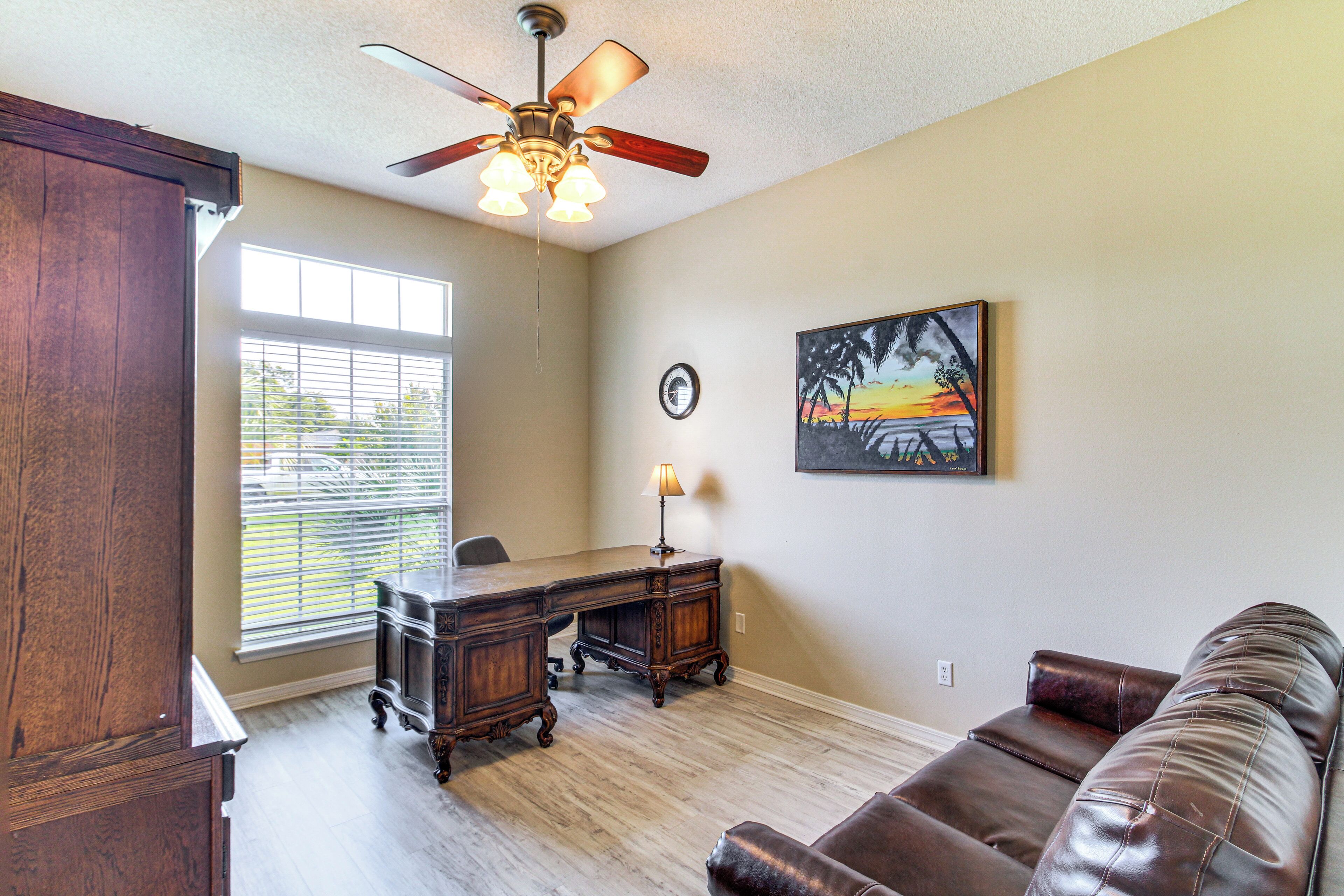 A room with a wooden desk, a leather couch, a ceiling fan, and a painting on the wall.