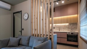 A modern living room with a grey sofa, a wooden partition, a kitchen with wooden cabinets, a built-in oven, and a wall clock. - Latitude 37 (Kalamata)