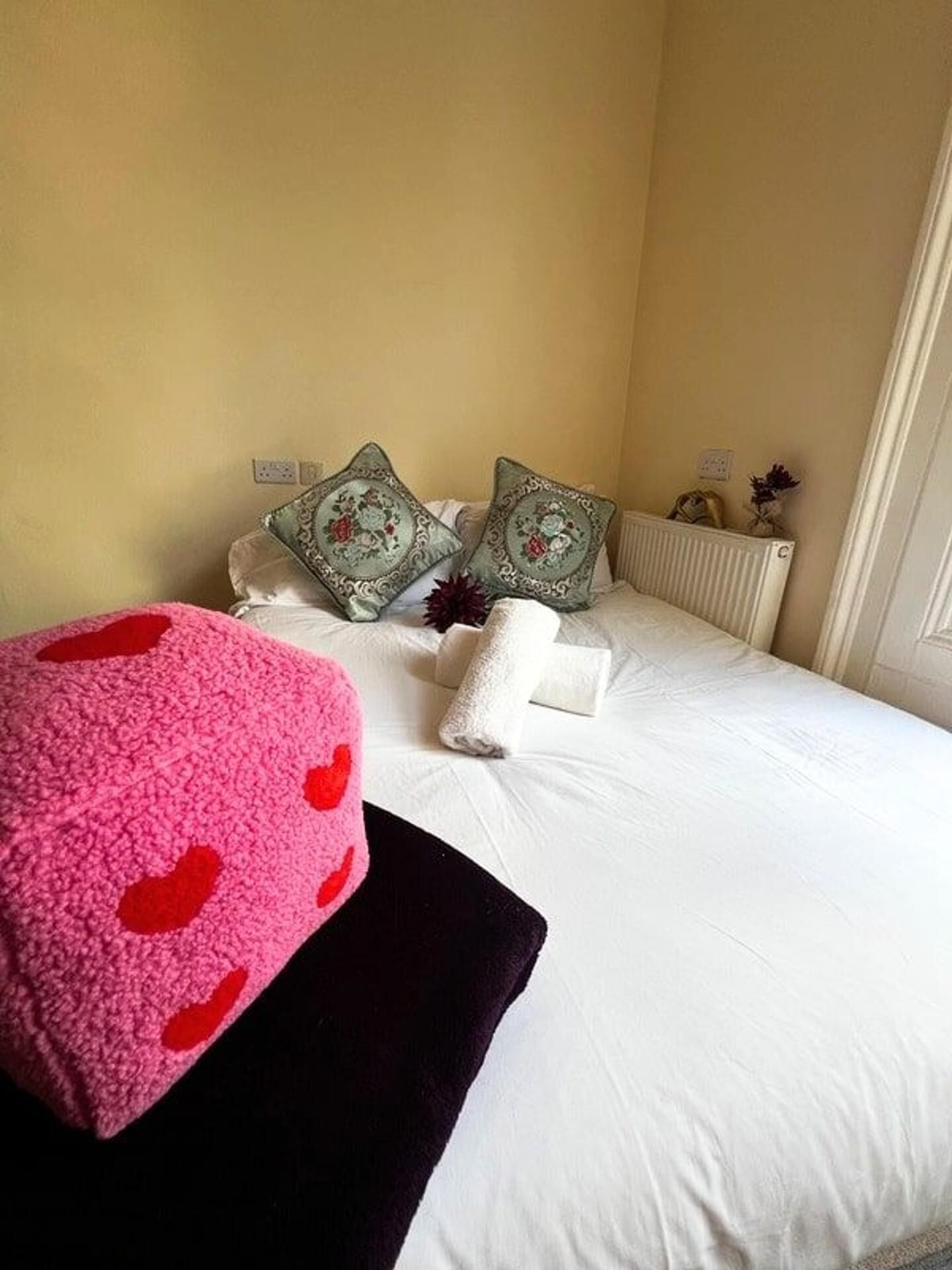 Una cama bien hecha con sábanas blancas y almohadones decorativos, un reposapiés rosa con estampado de corazones rojos y un radiador en la pared.
