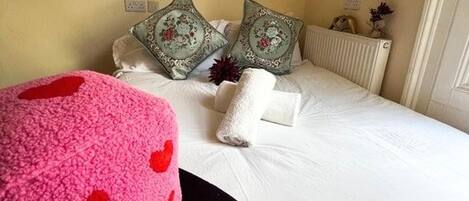 Una cama bien hecha con sábanas blancas y almohadones decorativos, un reposapiés rosa con estampado de corazones rojos y un radiador en la pared.