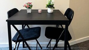 Una mesa pequeña de color oscuro con dos sillas, cada una con un cojín negro. Hay dos jarrones con flores sobre la mesa.