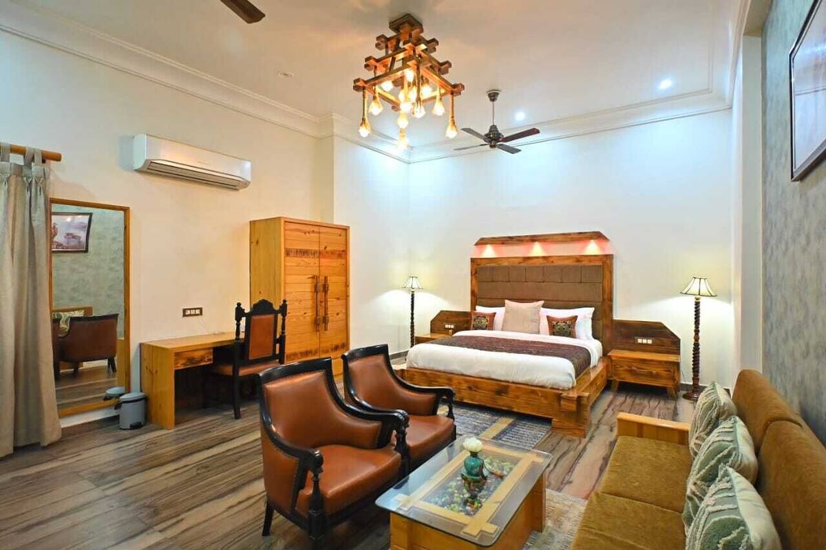 A hotel room with a large bed, two armchairs, a sofa, a wooden wardrobe, a desk, and a ceiling fan.