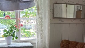 A room with a wooden floor, a grey dresser with drawers, a brown armchair, a white trash bin, and a window with lace curtains.
