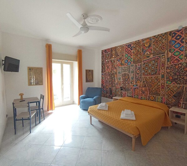 A hotel room with a bed, a desk, a chair, a blue sofa, a ceiling fan, and a patterned wall.