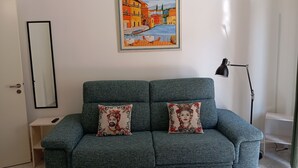 Un sofá color turquesa con cojines estampados, un cuadro enmarco de un pueblo costero y una lámpara de pie.