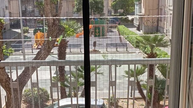 Vista a través de una ventana con una verja de metal, árboles y coches aparcados.