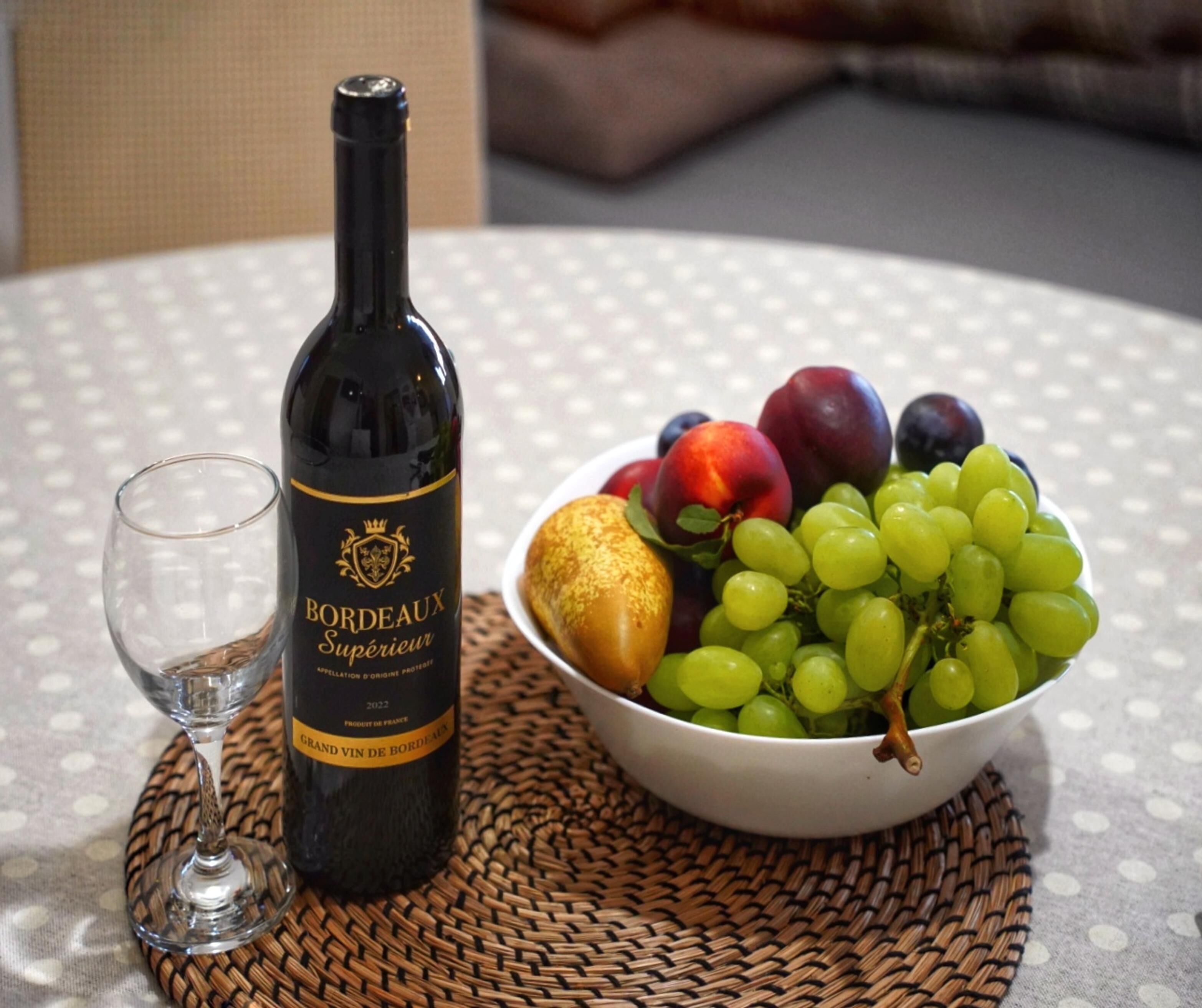 Una botella de vino Bordeaux Supérieur, una copa y un bol con uvas y ciruelas sobre una mesa.