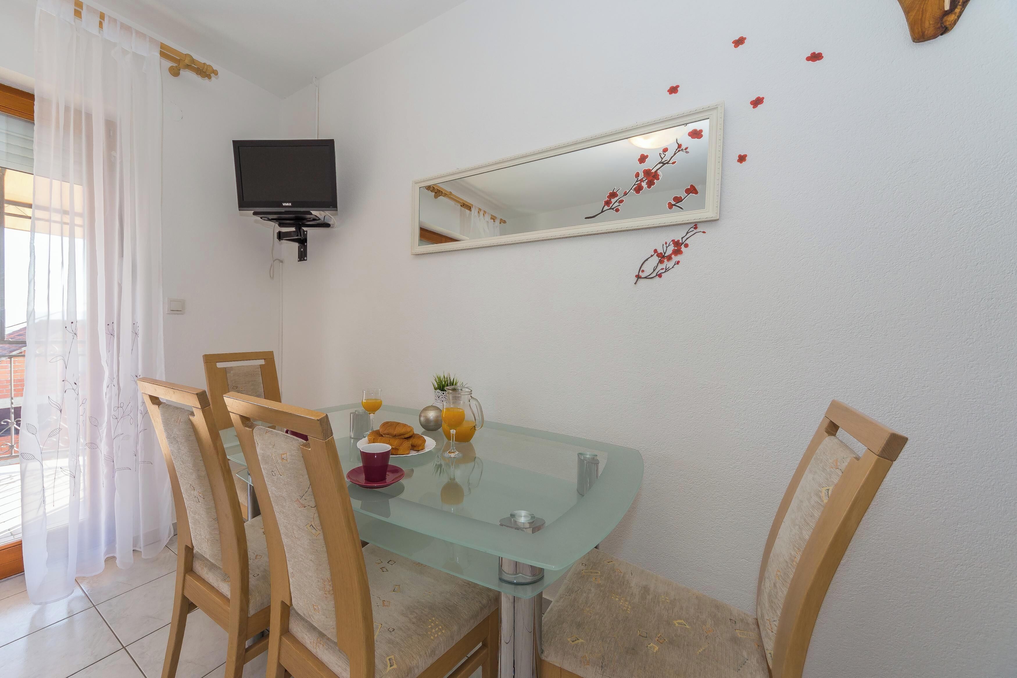 A dining area with a glass table, wooden chairs, a TV mounted on the wall, and a mirror with a floral design.