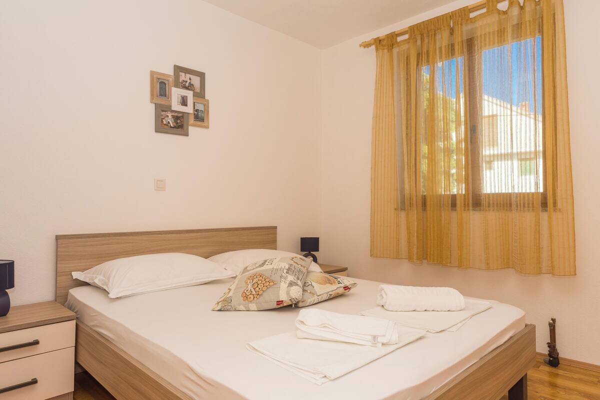 A neatly made bed with white linens and decorative pillows, a bedside table with a lamp, and a window with sheer curtains.