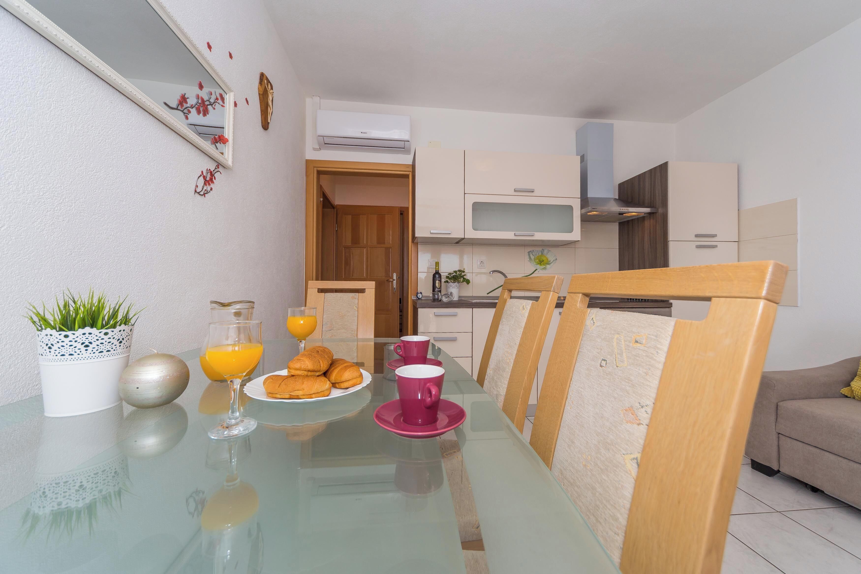 Une cuisine moderne avec des meubles blancs, une table à manger avec un plateau en verre et un coin petit-déjeuner comprenant des croissants et du jus d’orange.
