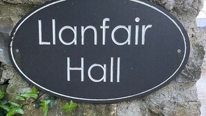 Een zwart bord met witte letters waarop 'Llanfair Hall' staat, bevestigd aan een stenen muur.
