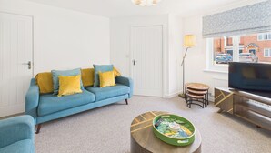 Un salon avec un canapé bleu, une petite table ronde sur laquelle est posé un plateau décoratif, un téléviseur sur un meuble en bois et une fenêtre donnant sur un bâtiment en briques rouges.