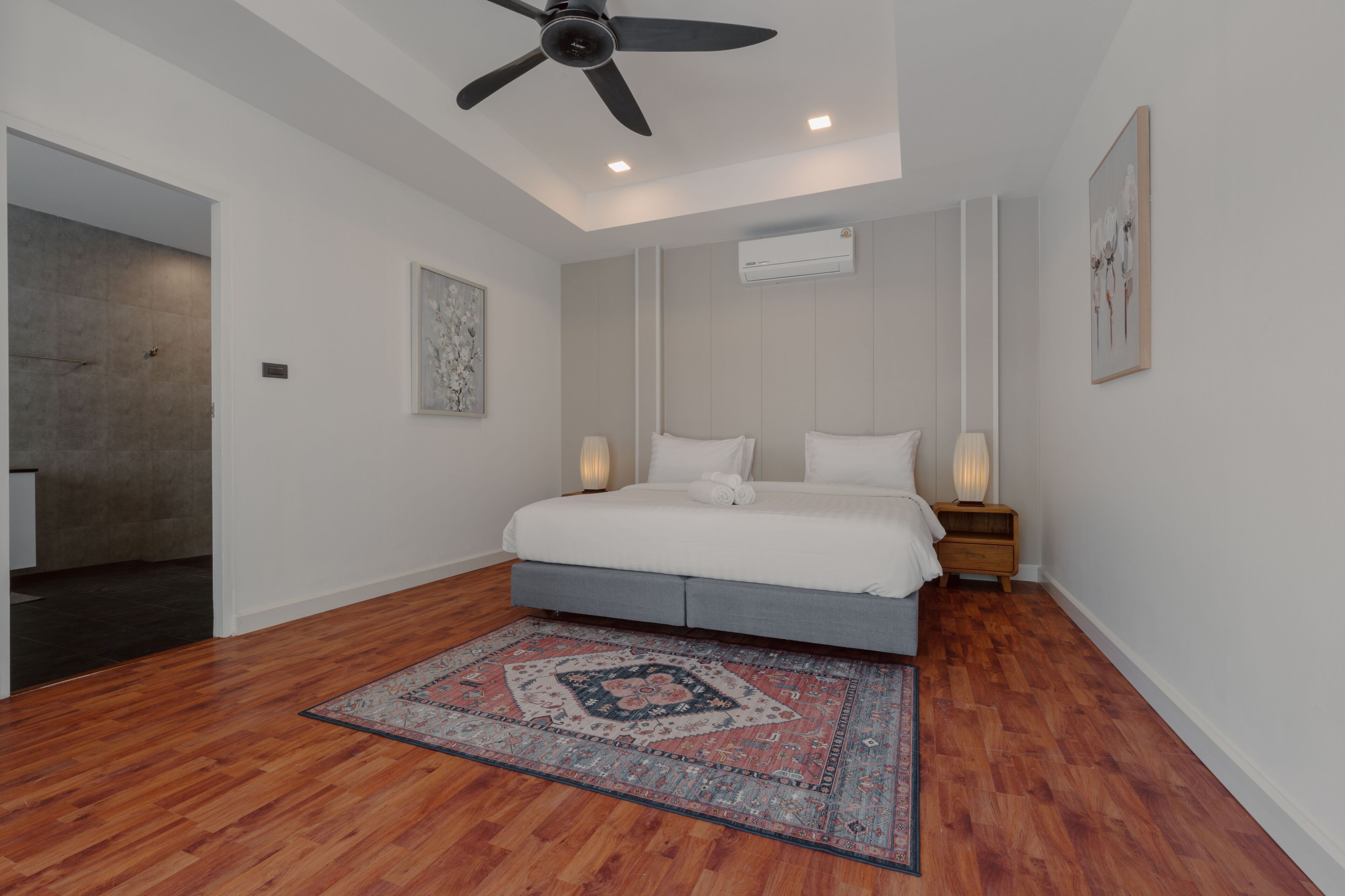 A bedroom with a bed, a ceiling fan, a wall-mounted air conditioner, and a patterned rug.