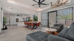 Una sala moderna con un sofá gris, un comedor con mesa y sillas, una cocina al fondo y un amplio ventanal corredero que da a un balcón.