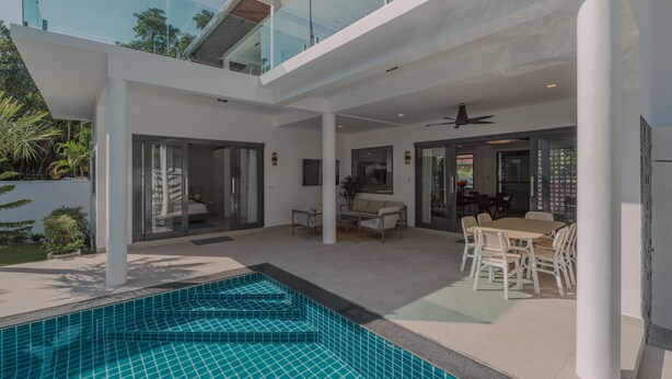 Un área de piscina exterior moderna con barandilla de vidrio, una zona de comedor con mobiliario blanco y vistas a la ciudad.