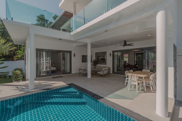 A modern outdoor pool area with a glass balustrade, a dining area with white furniture, and a view of the cityscape. - Happy Family Villa 3 (Mueng)