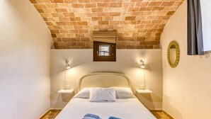 Un dormitorio con techo de ladrillo, una ventana pequeña, dos mesitas de noche, una cama con ropa de cama blanca y un espejo en la pared.