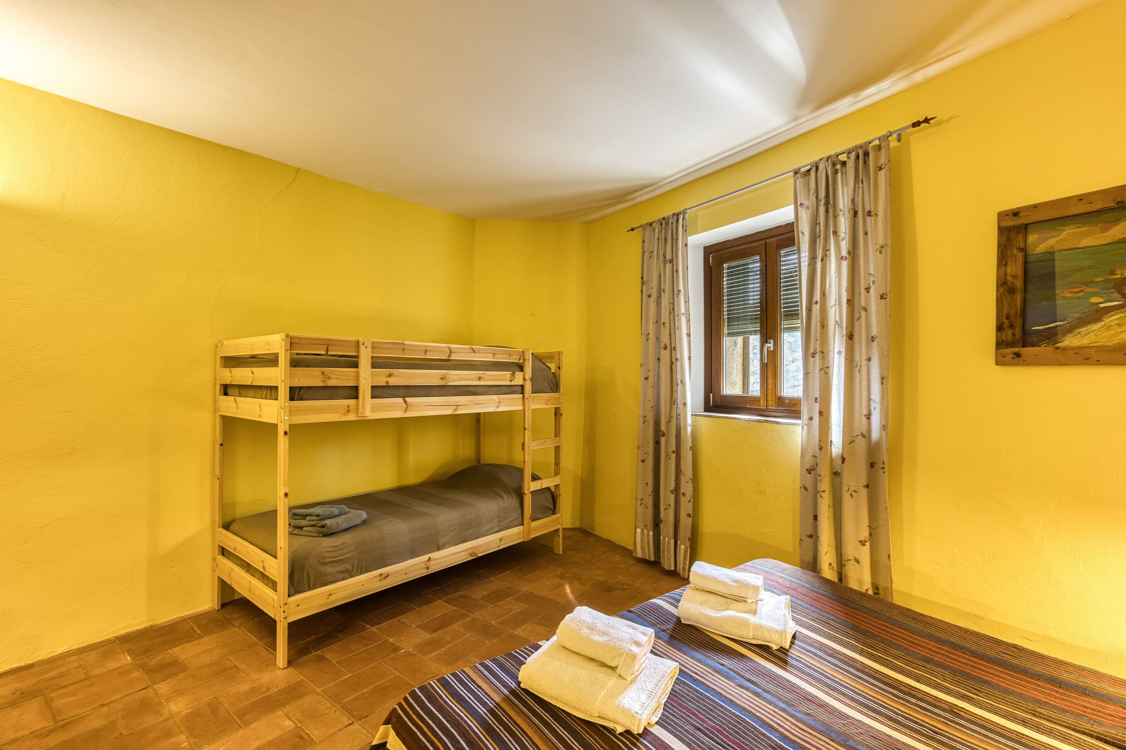 A room with a bunk bed, a window with curtains, a painting on the wall, and a striped rug on the floor.