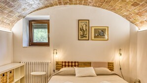 Une chambre avec un plafond en briques, une tête de lit en bois, une petite fenêtre, un radiateur blanc, une table de chevet blanche et deux tableaux encadrés accrochés au mur.