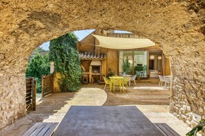 Um pátio externo com paredes de pedra, uma mesa de madeira, cadeiras amarelas e uma área de estar coberta.