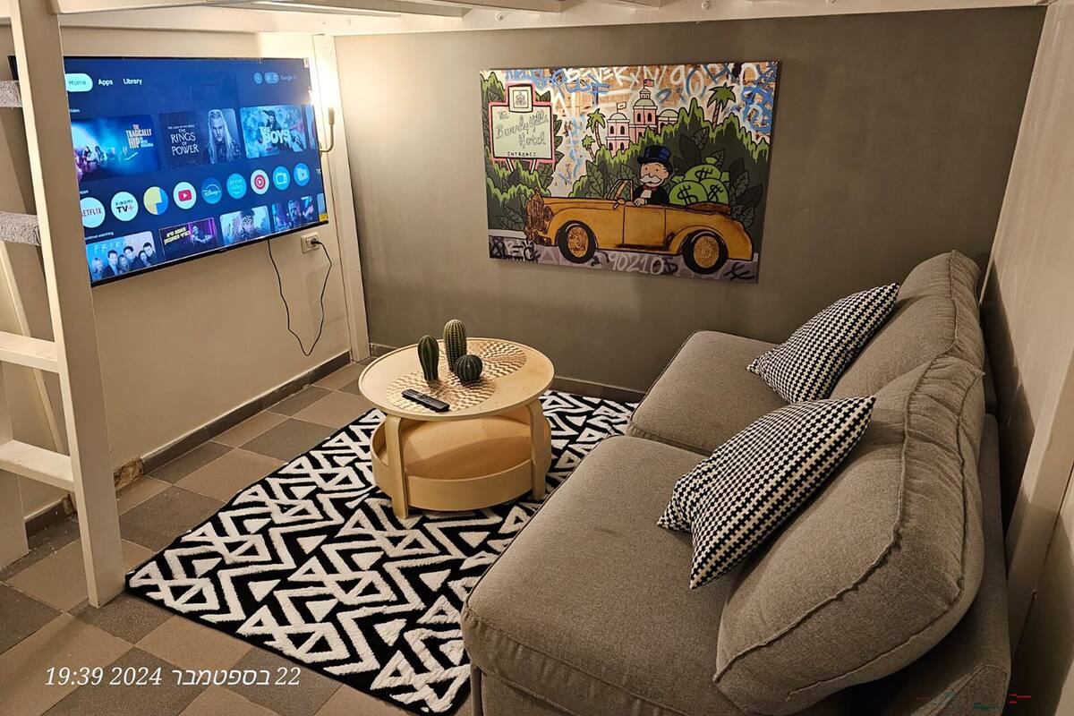 A modern living room with a grey sofa, black and white patterned rug, and a painting of a vintage car. A flat-screen TV is mounted on the wall displaying various streaming services.
