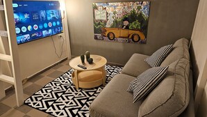 A modern living room with a grey sofa, black and white patterned rug, and a painting of a vintage car. A flat-screen TV is mounted on the wall displaying various streaming services. - SpaceART (Jerusalem)