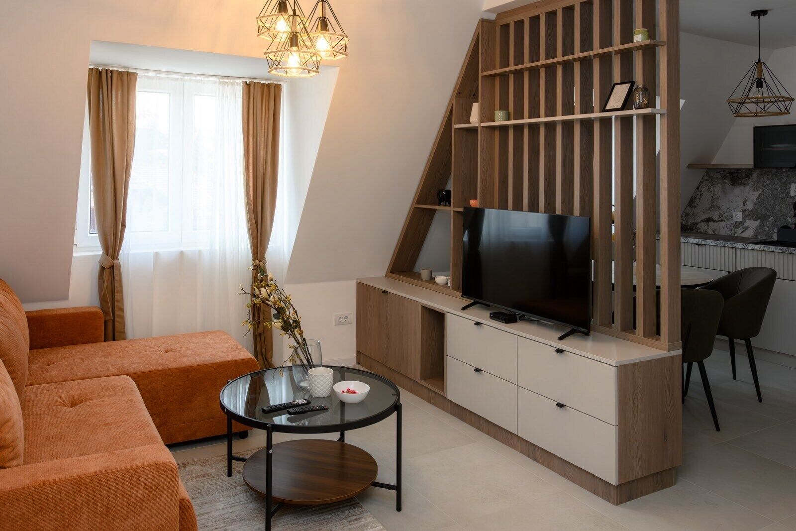 A modern living room with a sofa, a glass coffee table, a TV on a wooden cabinet, and a kitchen area with a dining table and chairs.