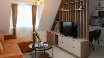 Una sala de estar moderna con un sofá, una mesa de centro de vidrio, un televisor sobre un mueble de madera y una zona de cocina con una mesa y sillas de comedor.