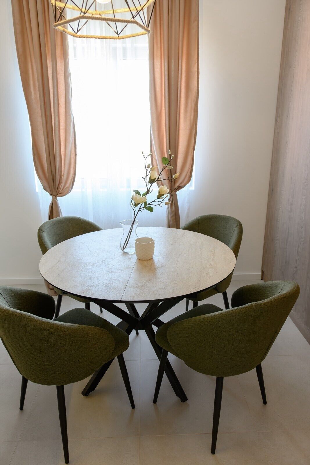 Een eethoek met een ronde tafel, zes groene stoelen, een vaas met witte bloemen en een hanglamp.