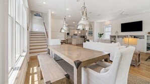 A modern kitchen with pendant lighting, a dining area with a wooden table and white chairs, and a living room with a sofa and a TV.