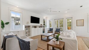 A spacious living room with a large TV, comfortable seating, and a view of the outdoors through large windows.
