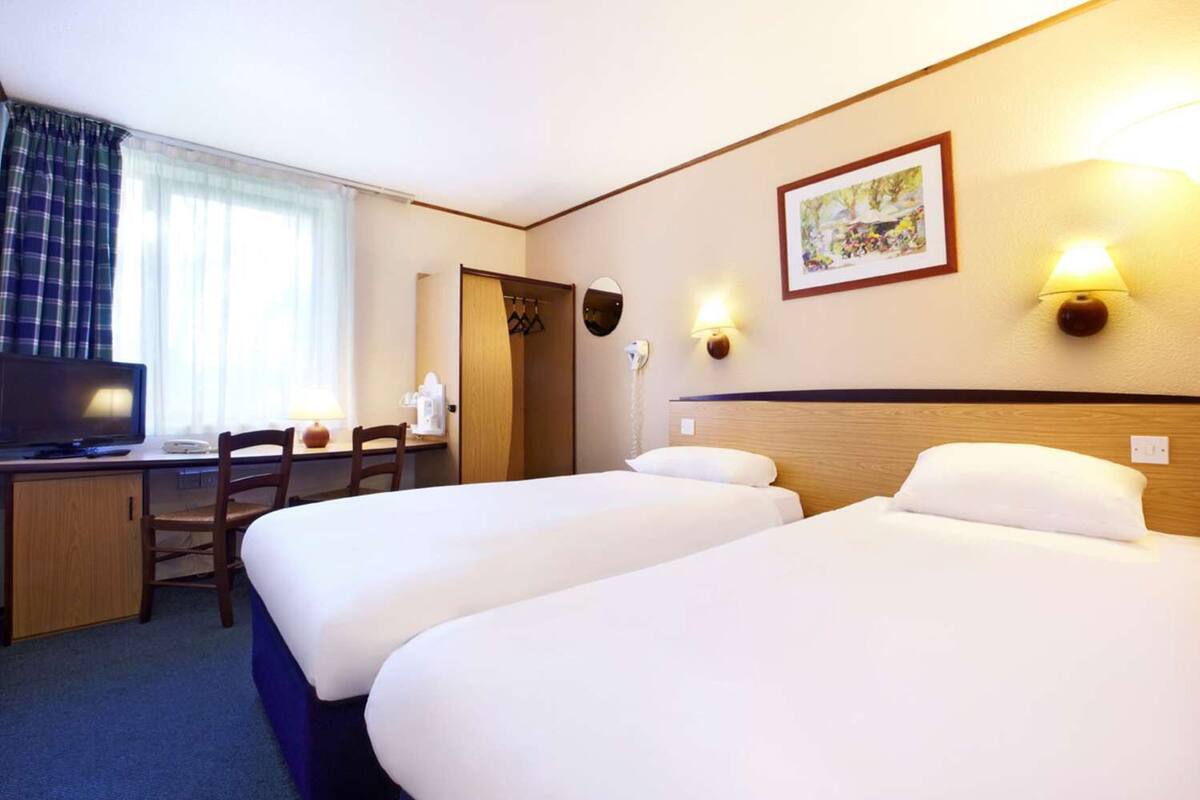 A hotel room with two beds, a desk with a computer, a chair, a lamp, and a painting on the wall.