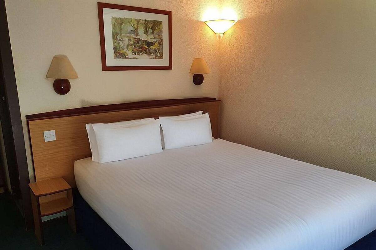 A neatly made bed with white linens, a wooden headboard, two bedside lamps, and a framed picture on the wall.