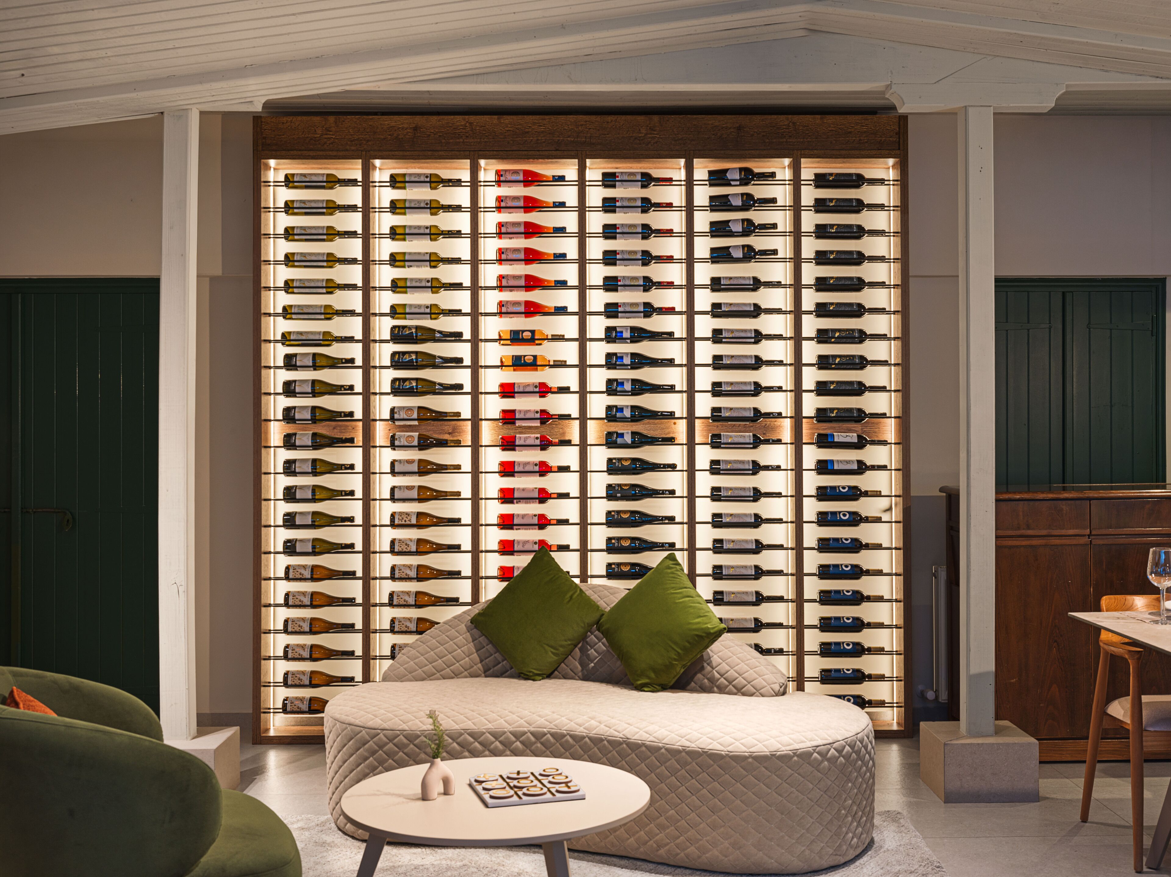 A room with a wall of wine bottles, a round seating area with cushions, and a small table with a book.