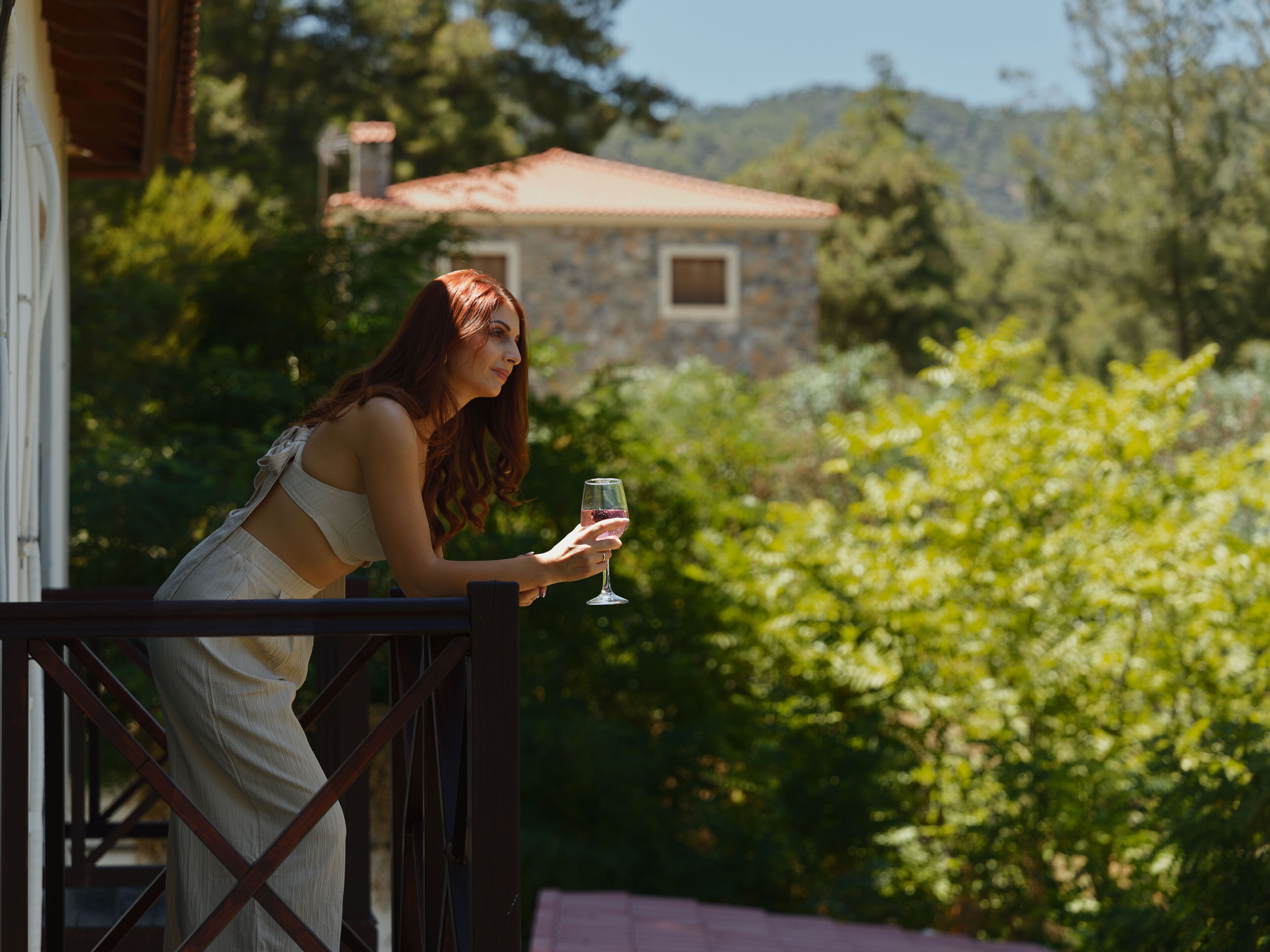 Una persona en un balcón sosteniendo un vaso, con vista a un jardín y un edificio al fondo.