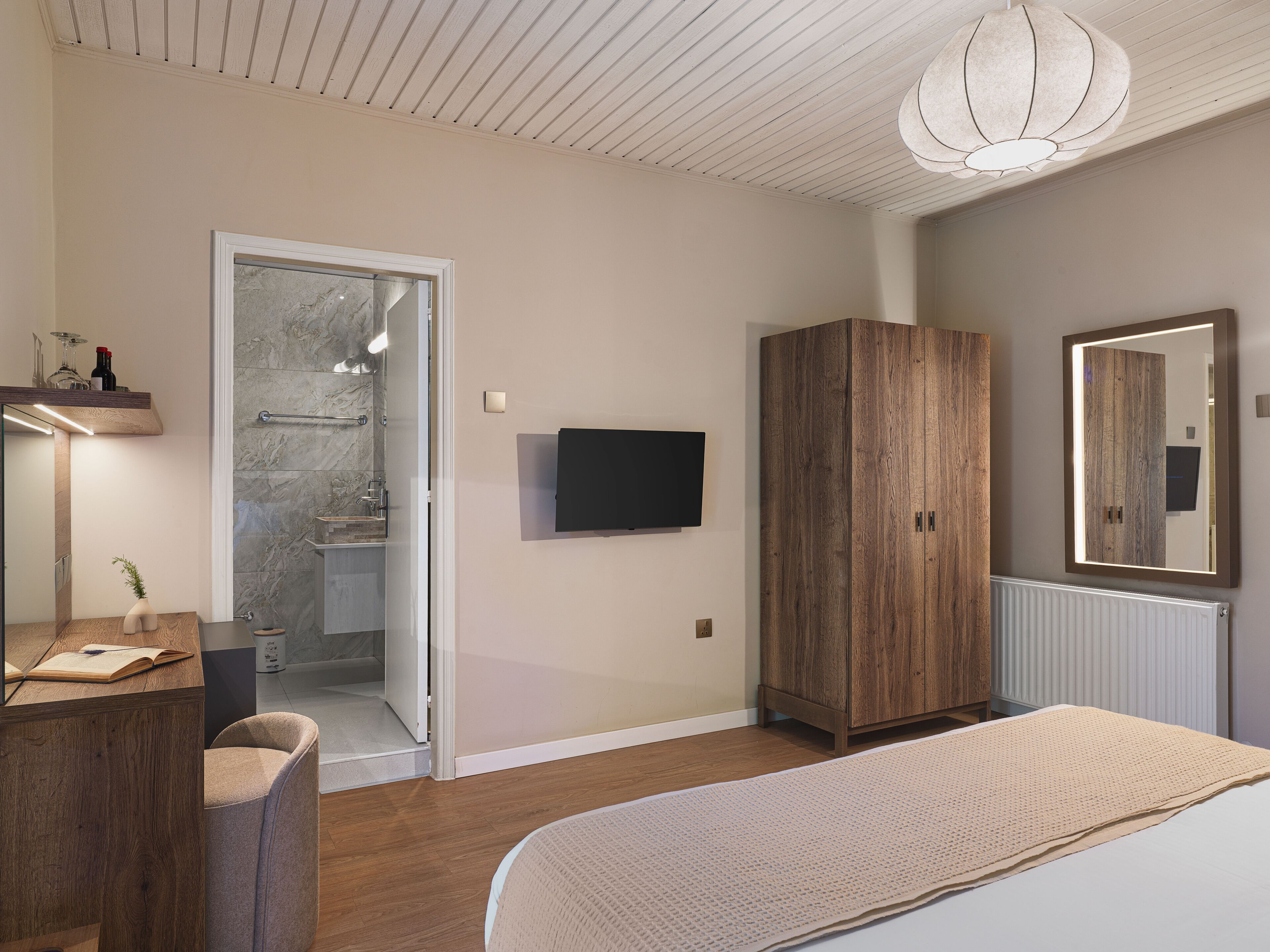 A bedroom with a bed, a wooden wardrobe, a television, a bathroom with a shower, and a desk with a chair.