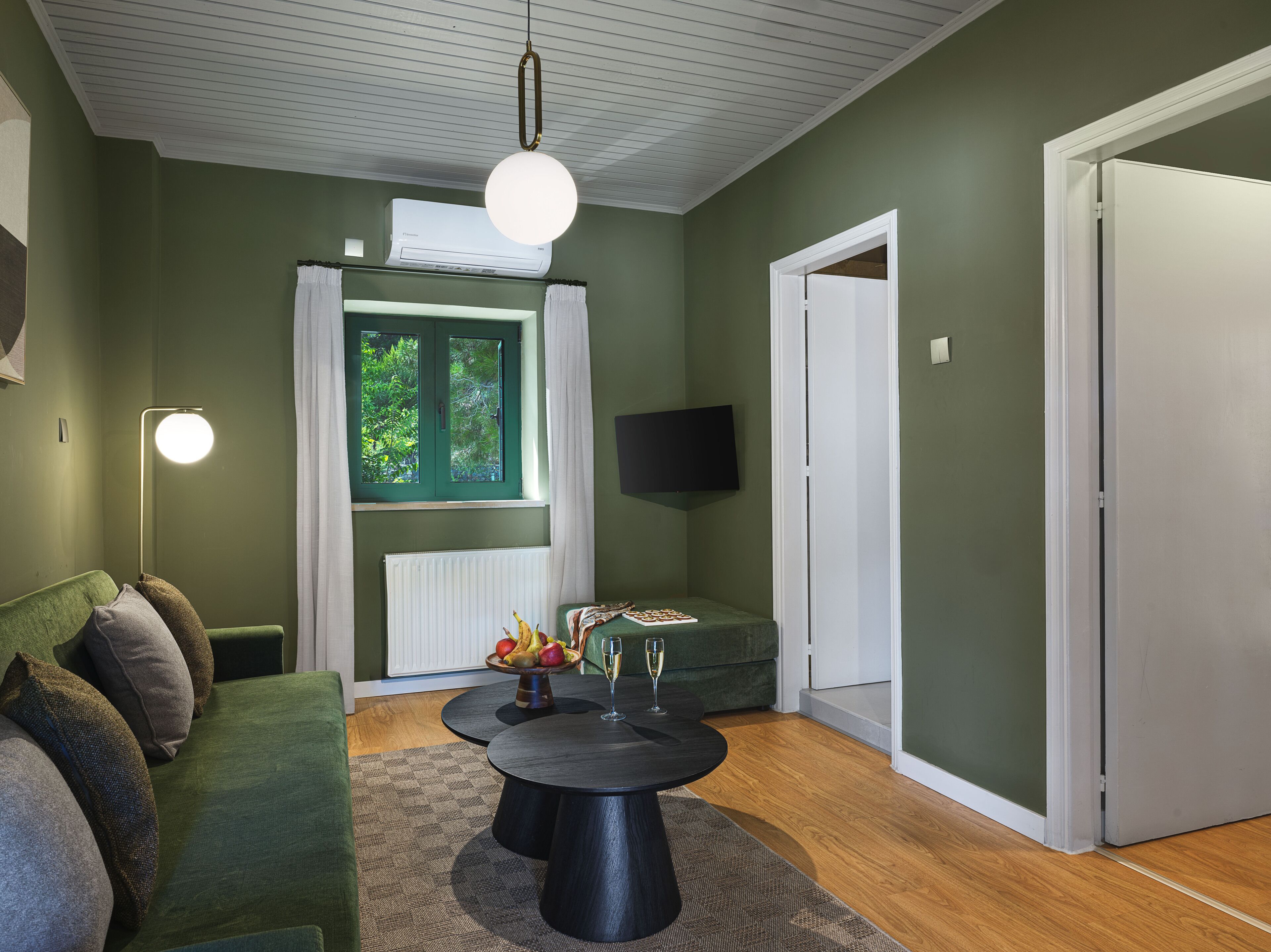 A living room with a green sofa, a round coffee table with fruit and a glass, a hanging light fixture, a window with curtains, and a flat-screen TV mounted on the wall.