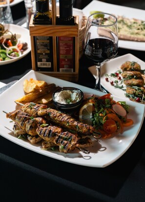 Un plato de brochetas de pollo a la parrilla, una copa de vino tinto y una botella de aceite de oliva sobre una mesa.
