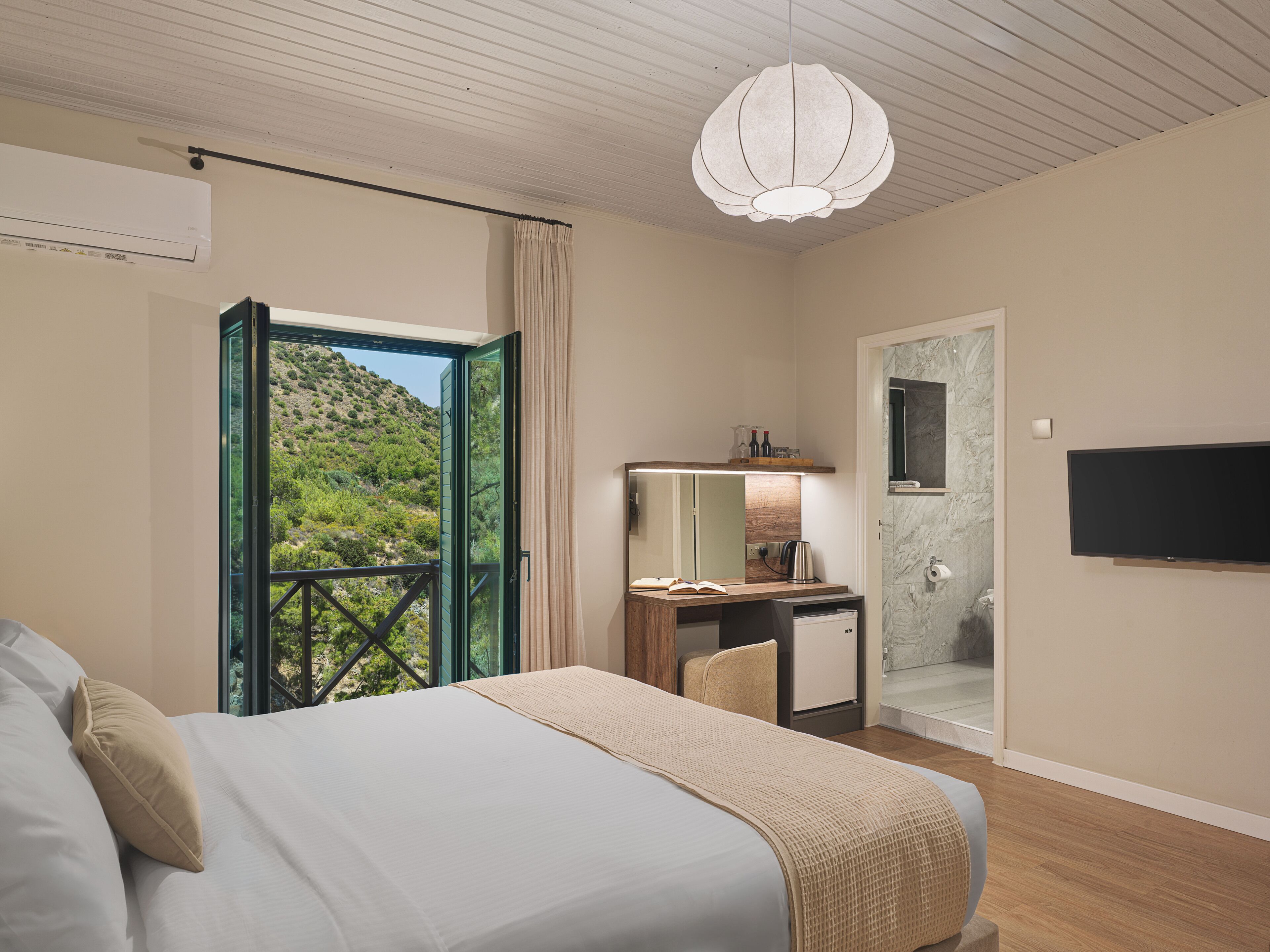 A hotel room with a bed, a desk, a TV, a bathroom, and a balcony with a view of greenery.