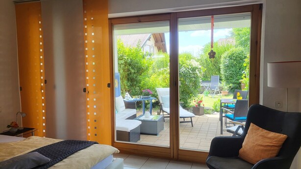 Ein Schlafzimmer mit einem Bett, einem Sessel, einer Glastür zu einer Terrasse und Blick auf einen Garten.