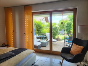 A bedroom with a bed, a chair, a sliding glass door to a patio, and a view of a garden.