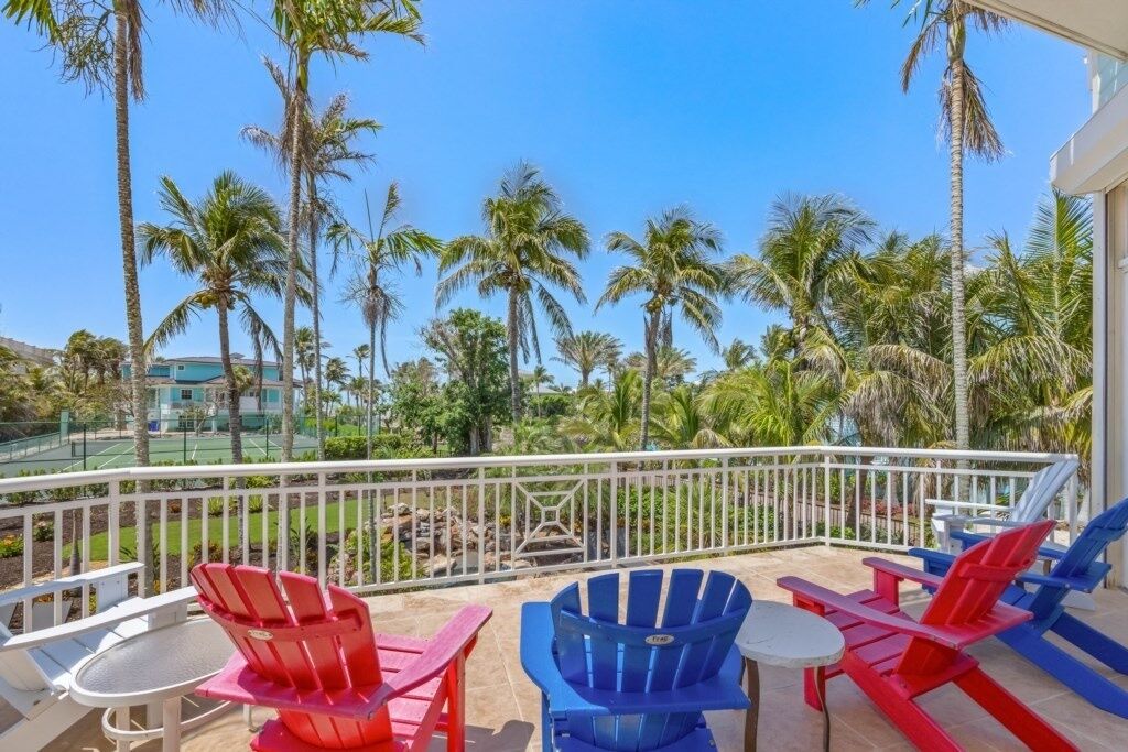 A balcony with red and blue Adirondack chairs, a small round table, and a view of palm trees and a tennis court.