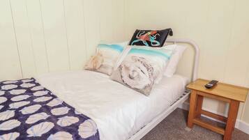 Una cama con estructura blanca, una colcha estampada y almohadas decorativas. Una mesita de noche de madera con un control remoto está al lado de la cama.