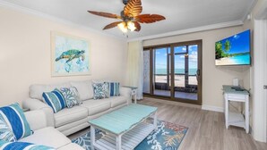 Sala de estar con sofá, mesa de centro y obras de arte. Hay un ventilador de techo, un televisor y vista a la playa a través de un ventanal.