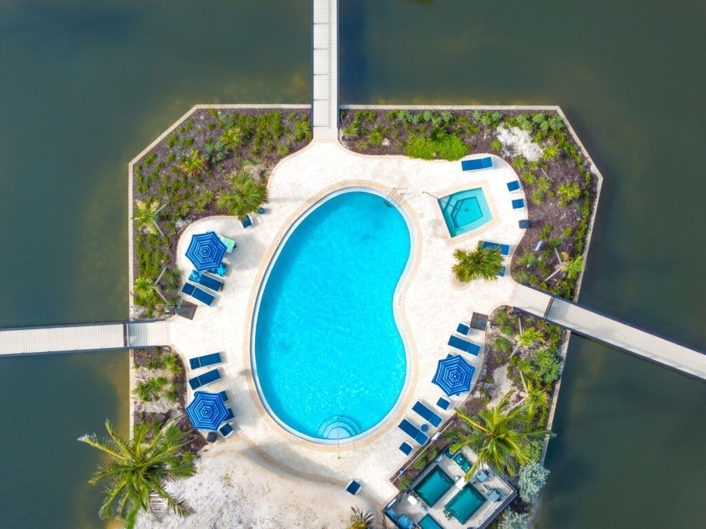 Vista aérea de un resort con piscina central rodeada de sillas de descanso y sombrillas, conectada al edificio principal mediante pasarelas.