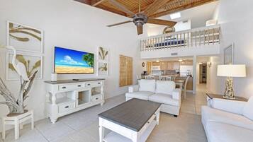 A spacious living area with a TV, white furniture, a ceiling fan, and a balcony.