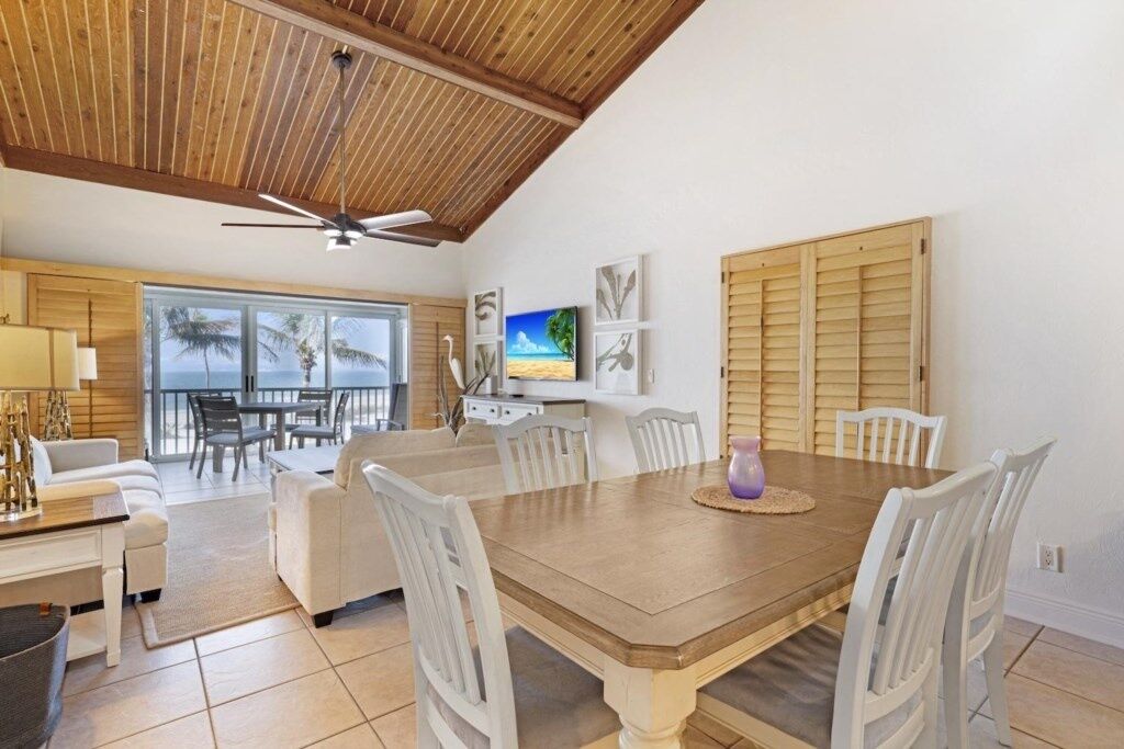 Un coin repas avec une table en bois et des chaises blanches, un ventilateur de plafond et une vue sur l’océan par de grandes fenêtres.