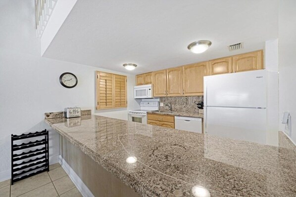 A kitchen with granite countertops, wooden cabinets, a white refrigerator, a microwave, and a clock on the wall.