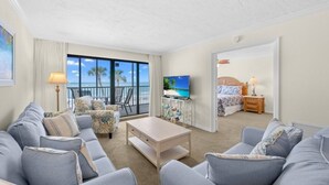 A spacious living room with a large window, a television, a sofa set, and a bedroom with a bed and nightstand. - Pointe Santo A25 2 Bedroom Condo (Sanibel)