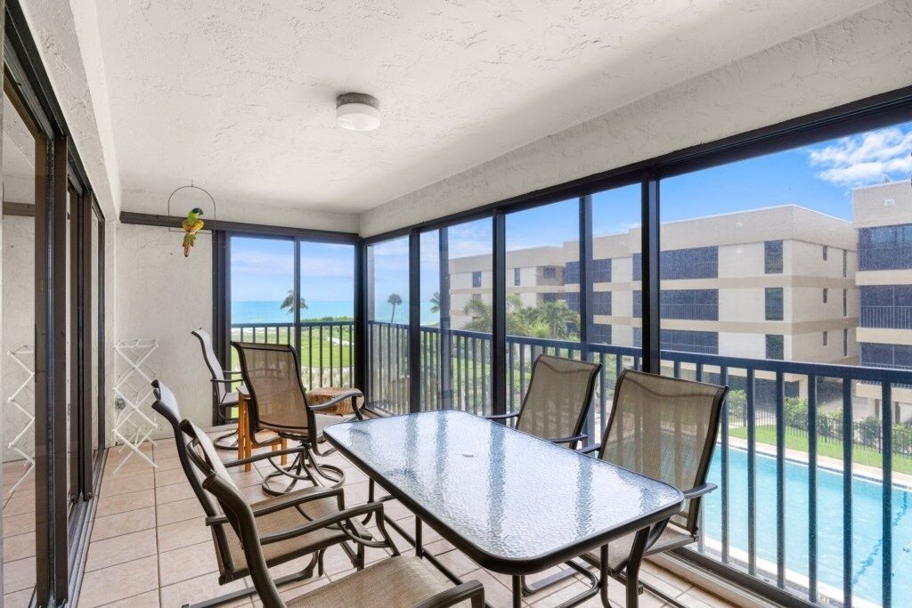A balcony with a glass table and chairs, a hanging bird feeder, and a view of a swimming pool and buildings.